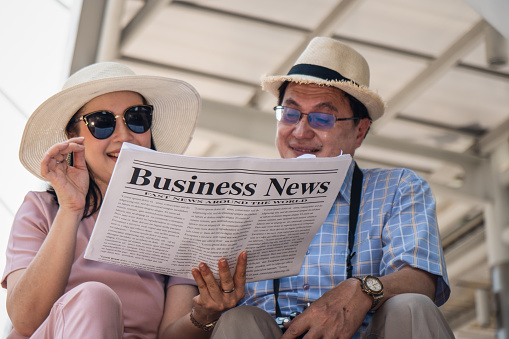 asian business traveler and tourist couple sitting on stairway reading business newspaper together