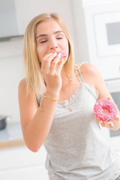 istock Young blonde girl eats pink donuts in home kitchen with taste emotions. 1159751281