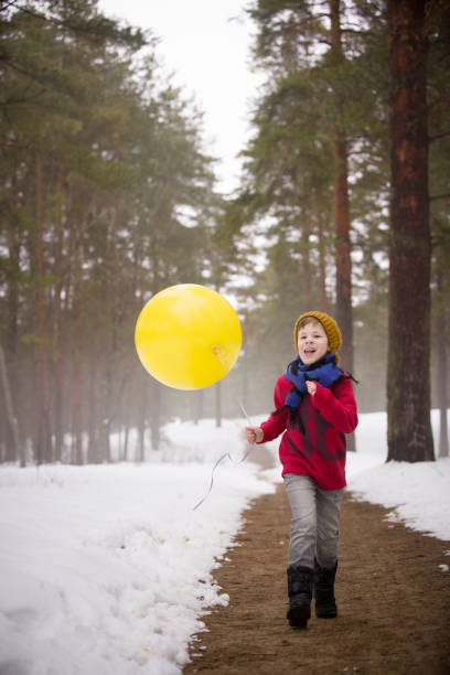 a small happy boy in jeans and a red sweater running in the winter forest and holding a big yellow balloon. birthday boy with balloon. birthday celebration. running boy with balloons. - balloon child winter snow imagens e fotografias de stock