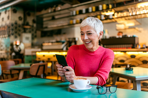 Photo of senior white hair businesswoman using a smartphone in a coffee shop during the day. Contemporary short hair senior woman with smartphone texting or reading the message while sitting in a cafe and smiling. Gorgeous female reading news via network on the mobile phone while waiting for her order in a modern coffee shop, European woman searching for information on cell telephone while sitting in the restaurant interior.