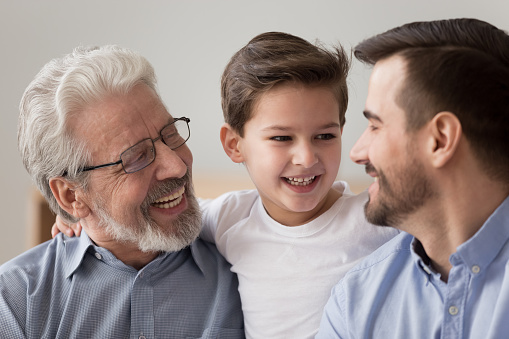 Close up of smiling preschooler boy child hug father and grandfather spend time together at home, happy three generations of men relax having fun laughing indoors, enjoy family weekend