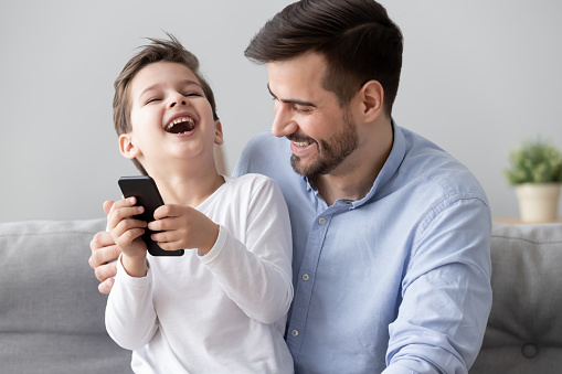 Close up of excited little boy hold smartphone having fun playing with young father at home, smiling happy dad and preschooler son relax on couch using cellphone enjoy leisure time spent together