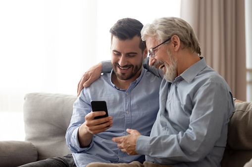 Excited elderly dad sit on couch hug grown son browsing internet on smartphone together, smiling millennial man and senior father have fun embrace enjoy leisure time at home using cellphone