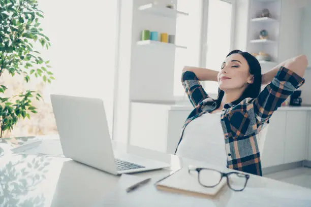 Photo of Portrait of cute charming entrepreneur touch head palms hands close eyes feel satisfied sit desk table big light apartment wear checkered shirt plaid modern