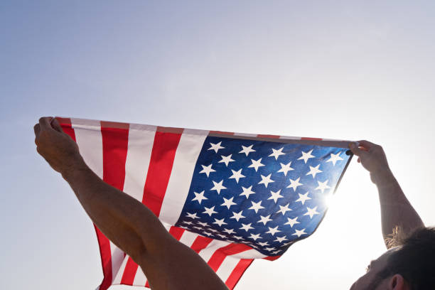 istock Man's raised hands with waving American Flag against clear blue sky 1159780590
