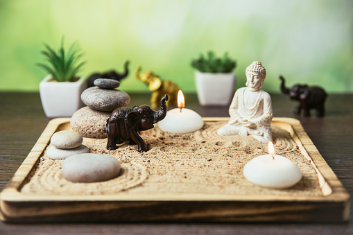 Miniature desk zen sandbox with Buddha figure sit in Lotus position, stacked zen sea stones, brown elephant figurines, spa candles burning against green bokeh studio background, copy space.