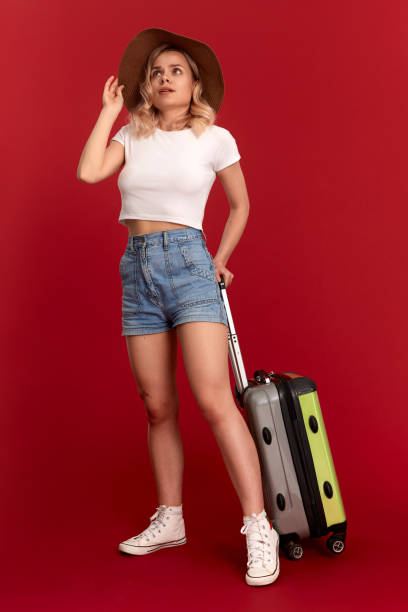 istock Happy young blond curly woman in a sundown hat holds grey luggage bag while standing infront of a red background 1159794565