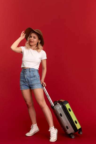 istock Happy young blond curly woman in a sundown hat holds grey luggage bag while standing infront of a red background 1159794574