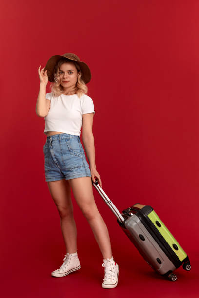 istock Happy young blond curly woman in a sundown hat holds grey luggage bag while standing infront of a red background 1159794580