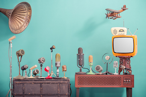 Retro old microphones for press conference or interview, outdated TV, radio, flying wooden toy plane and gramophone horn front gradient aquamarine wall background. Vintage old style filtered photo
