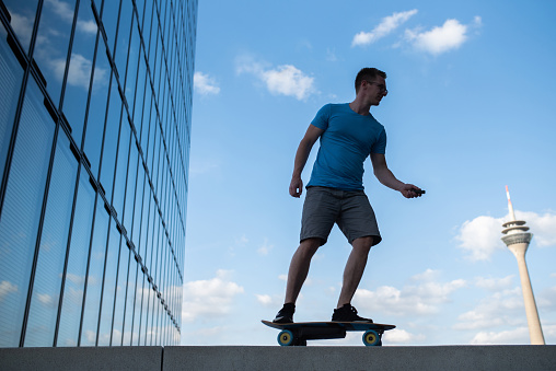 side view of a young male riding an electric skateboard in urban environment next to reflective windows of a big skyscraper at media harbor Duesseldorf with the rhine tower in the background
