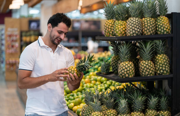 Young guy at the supermarket choosing a pineapple from shelf smiling - Healthy lifestyles