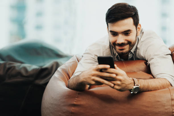 istock Smiling businessman using and relax smartphone device while sitting on sofa at home.  Man cheerful hipster guy typing an sms message at social network.  Technology Concept 1159835687