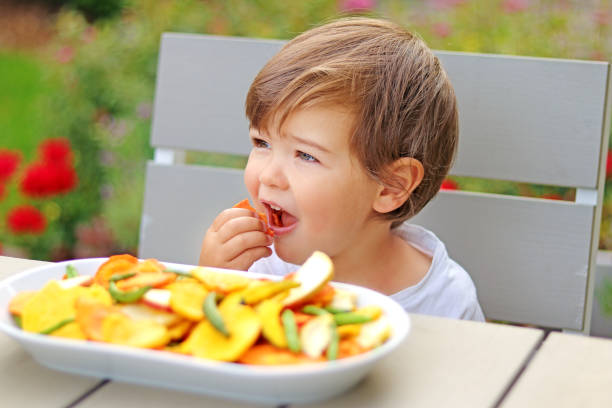 маленький малыш мальчик ест сушеные морковные овощи и фруктовые чипсы, сидя за столом в саду на открытом воздухе. натуральная здоровая орга - baby carrot snack healthy eating small стоковые фото и изображения
