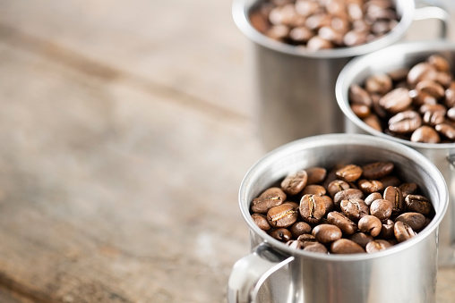 Three stainless steel cups full of dry roasted coffee beans. The cups have handles and are sitting on a wooden background.