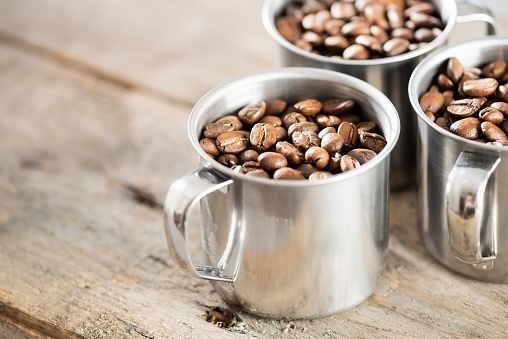 Three stainless steel cups full of dry roasted coffee beans. The cups have handles and are sitting on a wooden background.