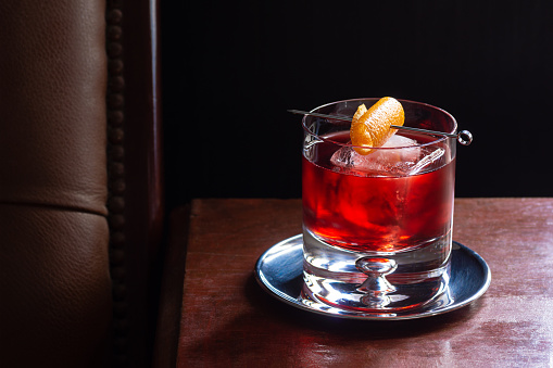 A Negroni cocktail, made from equal parts Campari, gin, and red vermouth in a rocks glass with ice and an orange twist. The drink is in a dark luxurious bar and there is copy space in the black background.