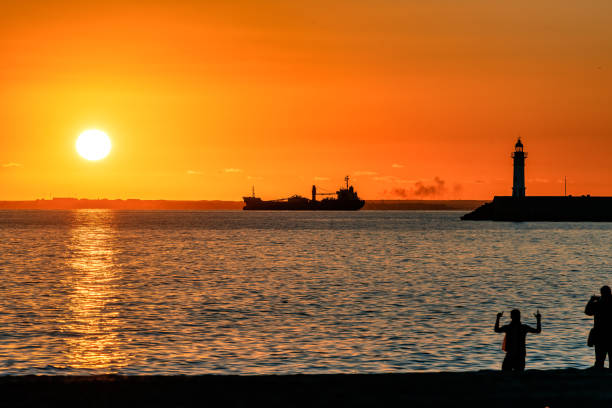 Red ocean sunset with silhouettes of cargo ship, lighthouse and people on the beach.