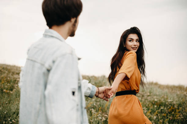 piękna urocza kobieta trzymająca za rękę swojego chłopaka prowadzącego go na pole z kwiatami w czasie wakacji podczas podróży. - couple old fashioned hipster holding hands zdjęcia i obrazy z banku zdjęć