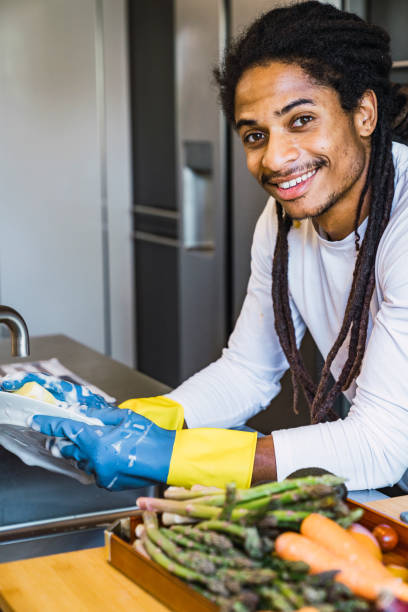Hand cleaning.Young black man washing dishes in modern kitchen.Young rastafarian woman washing dishes manually, by hand, wearing blue and yellow cleaning rubber gloves. Tired of cleaning, making a happy face. Looking top. limpieza manual. joven hombre negro lavando platos en la cocina moderna. joven mujer rastafari lavando los platos manualmente, a mano, usando guantes de goma de limpieza azul y amarillo. cansado de limpiar, hacer una cara feliz. mirando arriba.