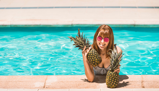 Caucasian redheaded girl with the body inside the water in the pool grabbed at the edge of the pool with two pine cones in her hands. Sticking out her tongue with sunglasses with red lenses.