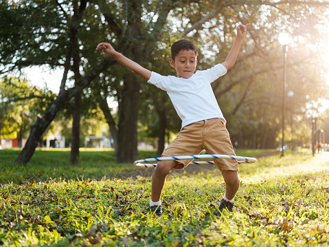 A horizontal photo of a boy in a park balancing a hula hoop while putting his arms up and with a concentrated expression.