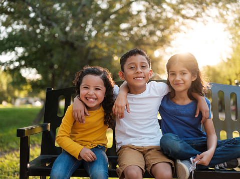 Three happy siblings sitting together outdoors on a park bench while smiling at the camera on a bright and sunny day
