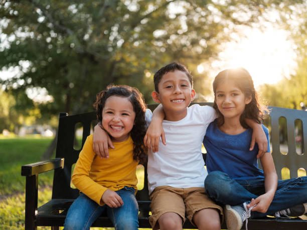 Happy siblings side side by side in the park Three happy siblings sitting together outdoors on a park bench while smiling at the camera on a bright and sunny day children only stock pictures, royalty-free photos & images