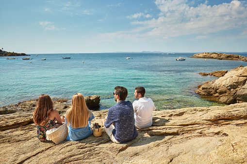 Young friends sitting together on rocks near Costa Brava water’s edge for a view of the Mediterranean Sea.