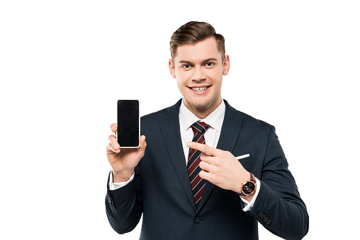 happy businessman in suit pointing with finger at smartphone with blank screen isolated on white