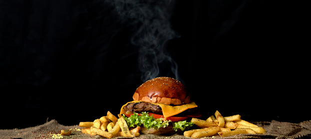 Giant delicious hot cheeseburger with tomato,lettuce,cheese and a big piece of meat served with french fries on a brown sack table mat on black background