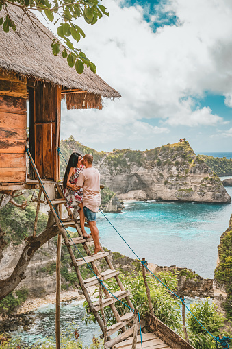 Photo of young couple kissing while standing on wooden stairs in Traditional house on a tree, looking at Atun beach, Nusa Penida island. Popular travel destination on Bali holidays. Indonesia. Kelingking Beach on Nusa Penida Island, Bali, Indonesia. Caucasian women and men an amazing wild nature view sandy beach with rocky mountains and azure lagoon with the clear water of the Indian ocean at sunset.