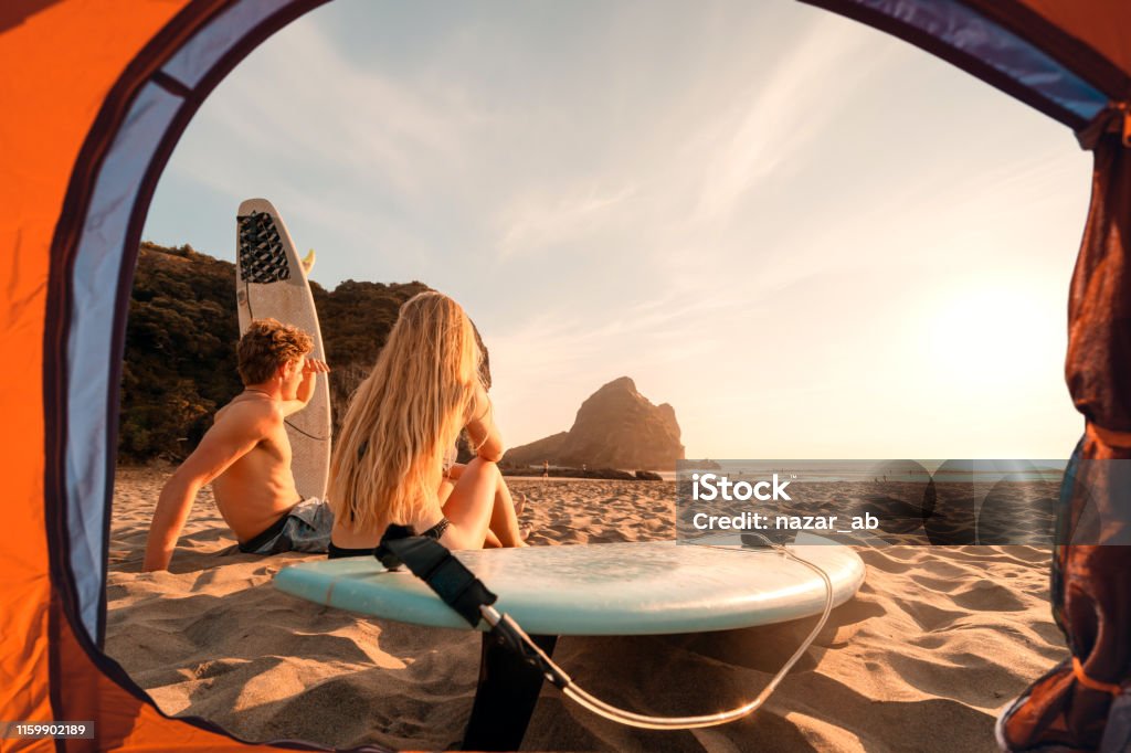 View of surfers from tent enjoying sunset at beach. Couple with surfboards enjoying sun going down in the evening. Beach Stock Photo