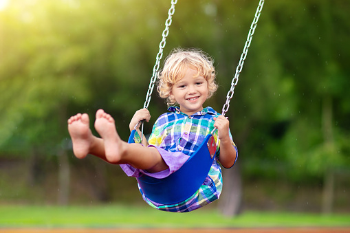 Child playing on outdoor playground in rain. Kids play on school or kindergarten yard. Active kid on colorful swing. Healthy summer activity for children in rainy weather. Little boy swinging.