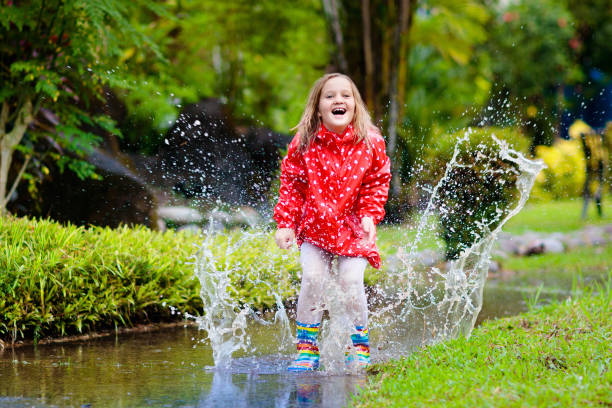 Child playing in puddle. Kids play and jump outdoor by autumn rain. Fall rainy weather outdoors activity for young children. Kid jumping in muddy puddles. Waterproof jacket and boots for little girl.