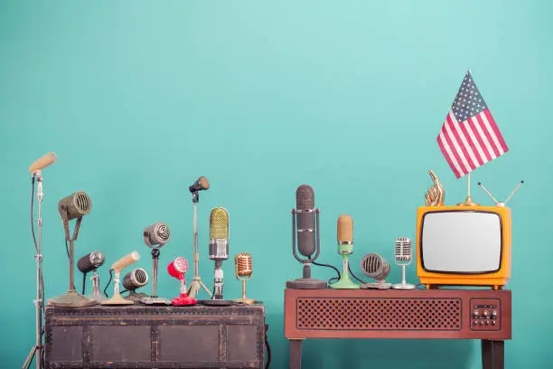 Photo of Retro old microphones from 50s and 60s for press conference or interview, outdated TV from 70s, USA flag, golden index finger front gradient aquamarine  wall background. Vintage style filtered photo