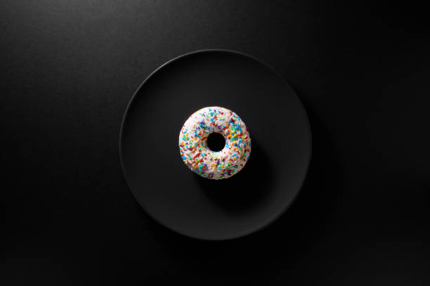 istock One donut with white icing served on black round plate on dark moody black background. 1159952173