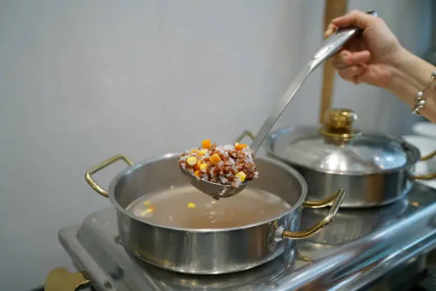 Photo of Homemade Soft Boiled Rice (Rice Porridge) with grains - Hand using dipper to ladle congee from hot pot, Traditional Chinese food style and Popular street food in Thailand.