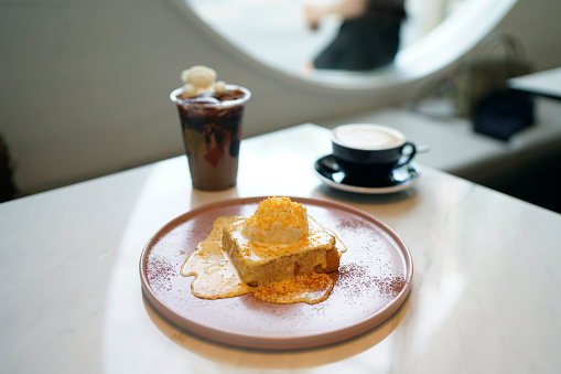 A plate of french toast (pain perdu) with salted yolk egg sauce, topped with vanilla ice cream and egg yolk flakes, served with hot latte and iced mocha coffee on marble table and blurred background.