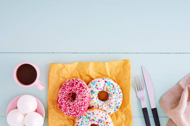 istock Colorful donuts on a paper bag with coffee cup, fork, knife and marshmallows on a pastel blue wooden table with copy space. Top view. 1159959707