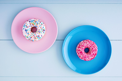 Two donuts. Pink donut is in turquoise plate and white doughnut is in pink plate on a pastel blue wooden table background. View from above.