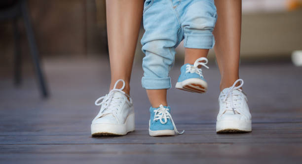 The first steps of the baby with the help of mom Mom in white sneakers with a young son, dressed in blue pants and blue shoes, a warm summer day and learn to walk in the street, the first steps, the mother maintains her son. first steps stock pictures, royalty-free photos & images