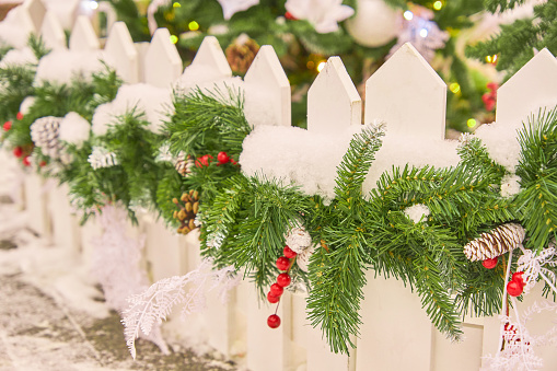 Close up of white fence decorated with christmas garland with cones and snow. Backyard festive decoration.