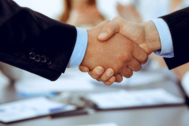 istock Business people shaking hands at meeting while theirs colleagues clapping and applausing. Group of unknown businessmen and women in modern white office. Success teamwork, partnership and handshake concept. 1159979305