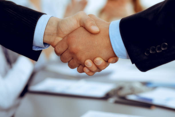 istock Business people shaking hands at meeting while theirs colleagues clapping and applausing. Group of unknown businessmen and women in modern white office. Success teamwork, partnership and handshake concept. 1159979309