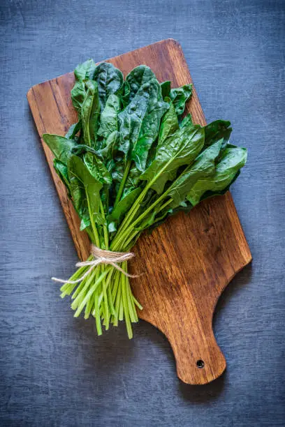 Top view of a rustic wooden cutting board with a spinach bunch on top on dark grey backdrop. Low key DSLR photo taken with Canon EOS 6D Mark II and Canon EF 24-105 mm f/4L