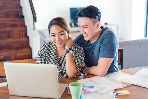 Young cheerful Asian couple sitting at the table at home and using laptop