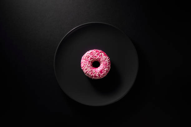 One donut with pink icing served on black round plate on dark moody black background.