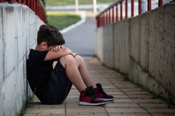 Photo of Little boy sad sitting alone at school hides his face. Isolation and bullying concept. Kid sad and unhappy, child was crying, upset, feel sick
