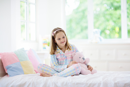 Child playing in bed in white sunny bedroom with window. Kids room and interior design. Little girl in pajamas at home. Bedding and textile for children nursery. Kid with toy. Nap and sleep time.